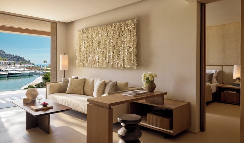 Two Bedroom Residence Balcony Marina Front iving area of a luxury hotel suite featuring a cream sofa, textured wall art, a wooden desk with a stool, and an open balcony overlooking a sunny yacht marina, with a glimpse into the bedroom at Sani Asterias by Kenwood Travel
