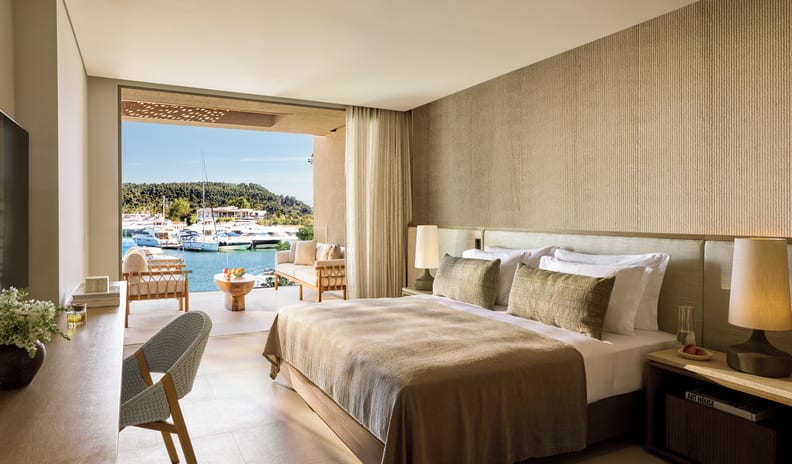Two Bedroom Residence Balcony Marina Front king-size bed, a wooden work desk, and an open sliding door to a private balcony overlooking a yacht marina at Sani Asterias by Kenwood Travel