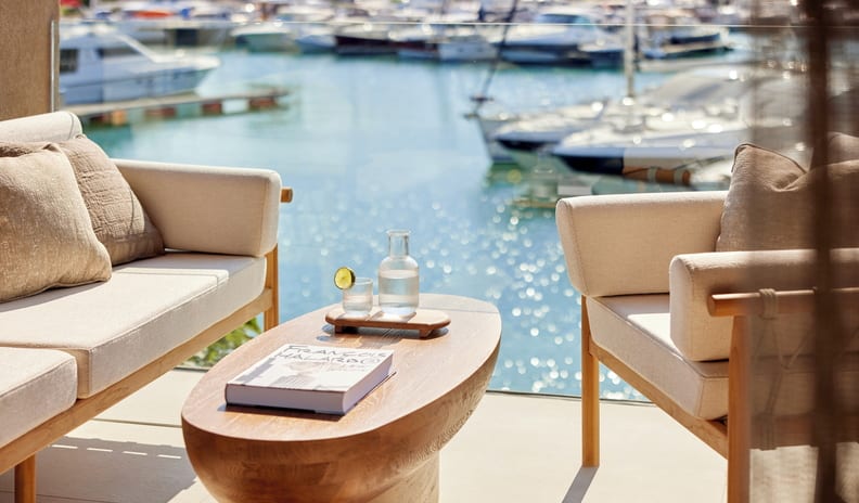 Two Bedroom Residence Balcony Marina Front balcony with stylish wooden outdoor sofas, an oval coffee table holding a drink with lime, and a sparkling blue marina full of yachts in the background at Sani Asterias by Kenwood Travel