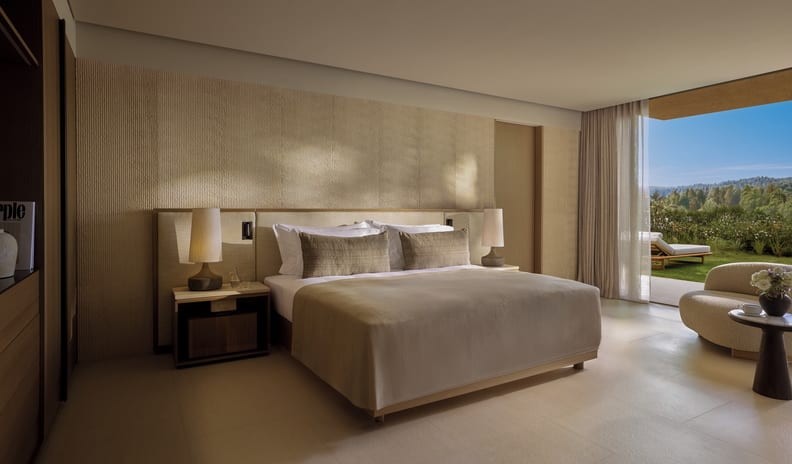 Two Bedroom Residence Pr. Garden Marina Front bedroom with a king-size bed, beige textured accent wall, and warm ambient lighting, featuring large sliding glass doors that open to a private lawn and garden view at Sani Asterias by Kenwood Travel