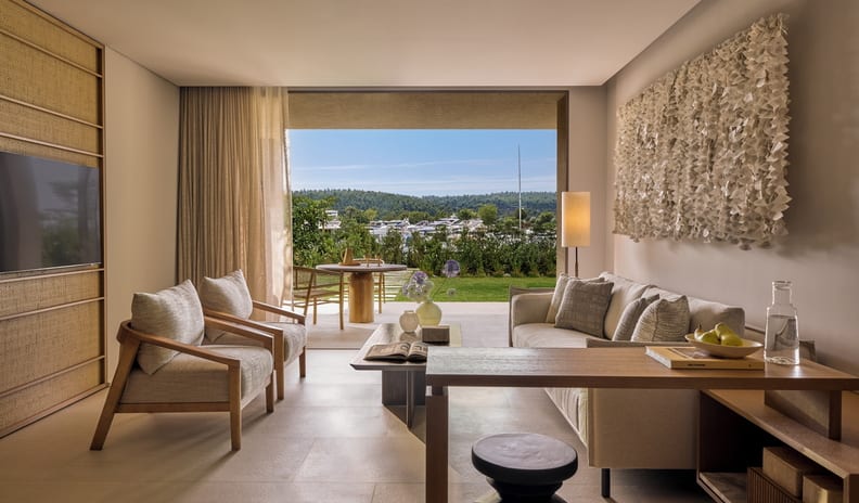 Two Bedroom Residence Pr. Garden Marina Fronttwo single beds with earth-toned linens, a ribbed beige accent wall, and an open glass door leading to a private patio with garden views at Sani Asterias by Kenwood Travel