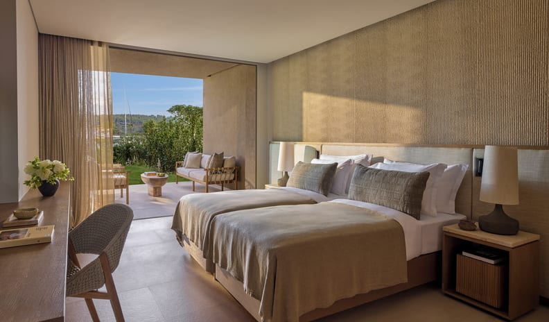 Two Bedroom Residence Pr. Garden Marina Front earth-toned bedding, a textured beige accent wall, and glass sliding doors leading to a private outdoor patio with garden views at Sani Asterias by Kenwood Travel