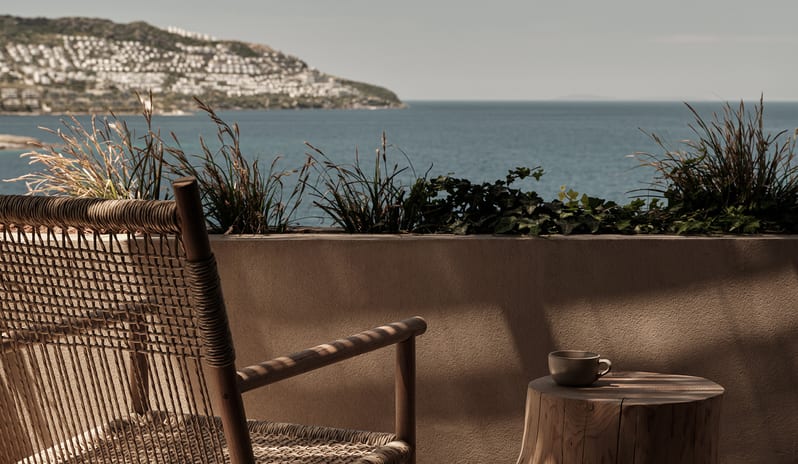 Classic woven wooden armchair, a natural tree stump side table with a coffee cup, and panoramic views of the Aegean Sea at OKU Bodrum by Kenwood Travel