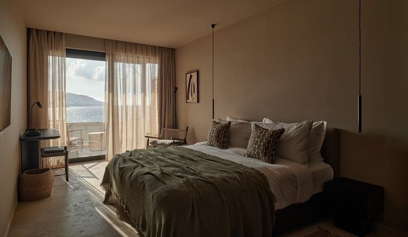 Classic king bed with olive green throw, minimalist decor, and large glass doors opening to a private balcony with Mediterranean sea views at OKU Bodrum by Kenwood Travel