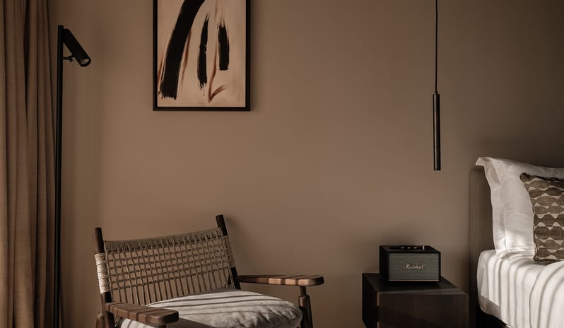 Classic bedroom reading nook showcasing a dark wood woven armchair, mid-century modern black pendant light, and minimalist framed abstract art on a beige wall at OKU Bodrum by Kenwood Travel