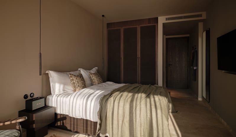 Classic bedroom design with earthy beige walls, sleek hanging pendant lights, a dark wood nightstand with a Marshall speaker, and patterned accent pillows at OKU Bodrum by Kenwood Travel