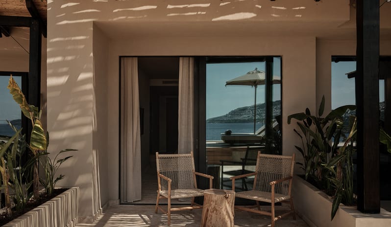 Classic Room with Shared Pool featuring two woven chairs, a rustic log side table, green tropical plants, and dramatic slatted shadows cast from a bamboo pergola overhead at OKU Bodrum by Kenwood Travel