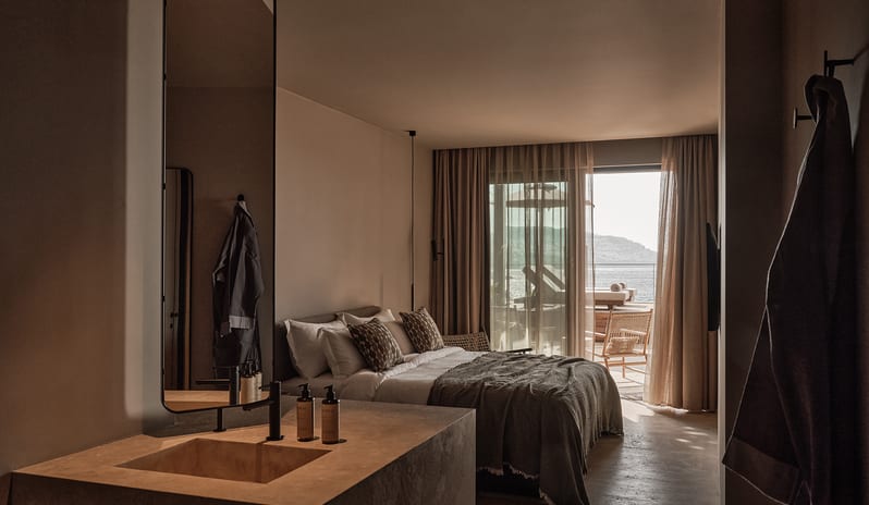 Classic Room with Shared Pool stone bathroom vanity sink with a tall mirror in the foreground, looking past a minimalist bed toward a sunlit private balcony with sea views at OKU Bodrum by Kenwood Travel