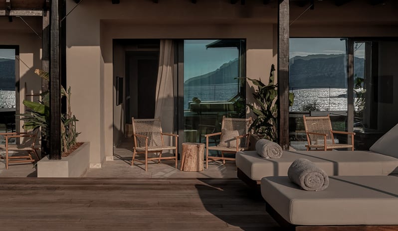 Classic Room with Shared Pool featuring a wooden pool deck with sun loungers, rolled towels, a private terrace with woven chairs, and a glass sliding door reflecting the sea and mountains at OKU Bodrum by Kenwood Travel