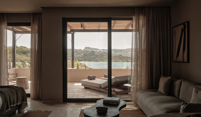 Deluxe Suite earth-toned sofa, a curved dark wood coffee table, and expansive sliding glass doors that open to a private terrace with sun loungers and a stunning view of the turquoise sea and coastline at OKU Bodrum by Kenwood Travel
