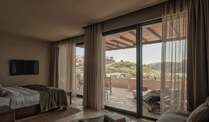 Deluxe Suite minimalist interior design, a large bed, organic wood coffee table, and floor-to-ceiling sliding glass doors opening onto a private sun terrace with hill views at OKU Bodrum by Kenwood Travel