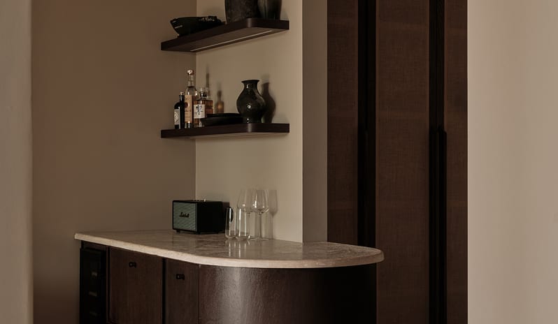 Junior Suite home bar setup with a curved dark oak cabinet, textured stone countertop, under-shelf lighting, and earth-toned ceramic decor at OKU Bodrum by Kenwood Travel