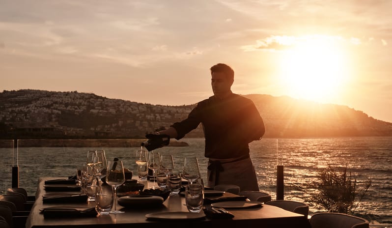 Teyo A waiter pouring wine at an outdoor dining table during a golden sunset over the sea at OKU Bodrum by Kenwood Travel