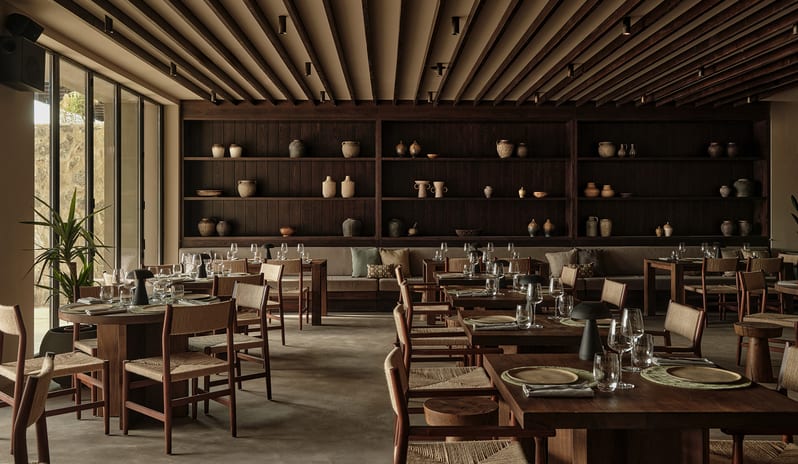 To Kima dark wood ceiling beams, rustic wooden tables, woven-seat chairs, and a large display shelf filled with artisanal pottery at OKU Bodrum by Kenwood Travel