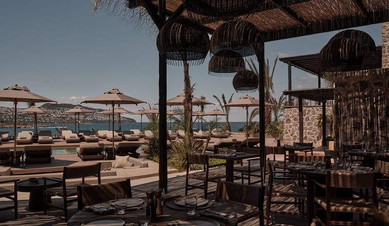 To Kima dark wood tables, woven chairs, and large spherical wicker pendant lights under a thatched pergola casting slatted shadows at OKU Bodrum by Kenwood Travel