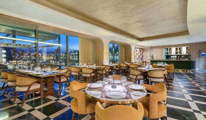 Riviera Modern restaurant interior featuring cane-back chairs, yellow armchairs, a green bar counter, and a black and white checkered marble tile floor at Rixos Radamis Sharm El Sheikh by Kenwood Travel