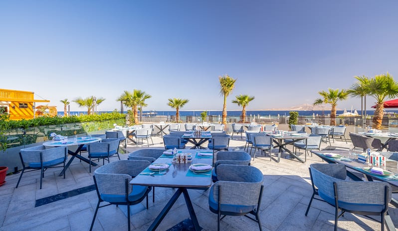 Salt beachfront resort dining patio showcasing beautifully arranged tables, blue armchairs, and scenic coastal views framed by palm trees at Rixos Radamis Sharm El Sheikh by Kenwood Travel