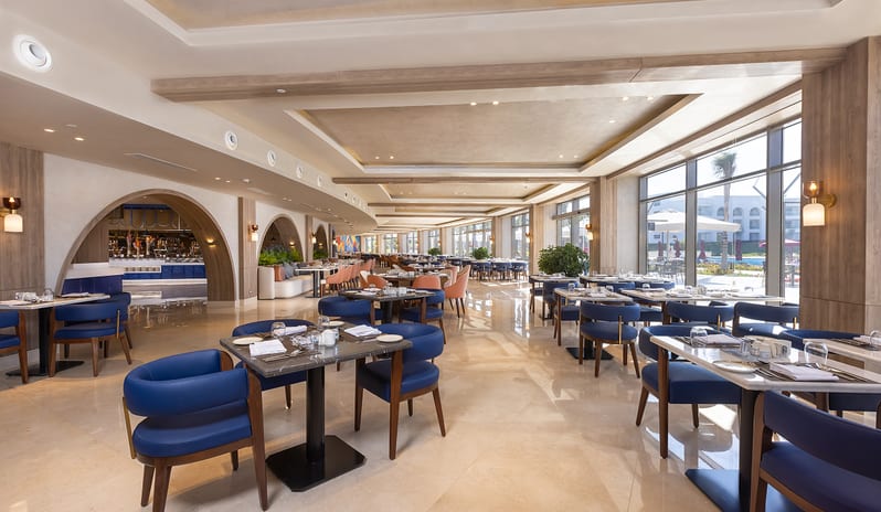 Turquoise large, brightly lit resort dining area with rows of neatly set tables, blue upholstered armchairs, wooden archways, and a view of an outdoor pool area at Rixos Radamis Sharm El Sheikh by Kenwood Travel
