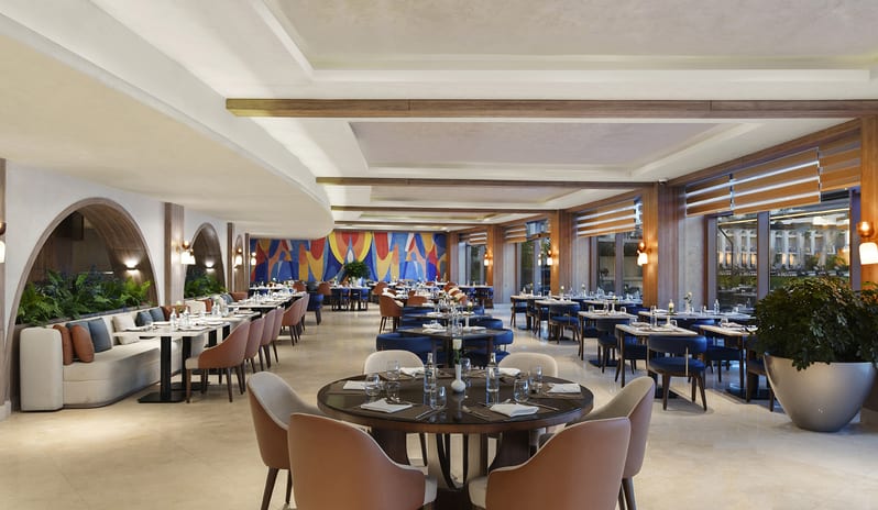Turquoise large, brightly lit resort dining area with rows of neatly set tables, blue upholstered armchairs, wooden archways, and a view of an outdoor pool area at Rixos Radamis Sharm El Sheikh by Kenwood Travel