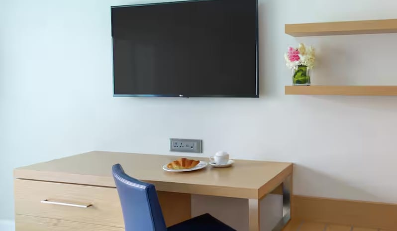 Island View Doubles wooden hotel room work desk with a blue leather chair, a flat-screen TV mounted on the wall above, and room service breakfast featuring a croissant and coffee at Harbor Club St. Lucia, Curio Collection by Hilton by Kenwood Travel
