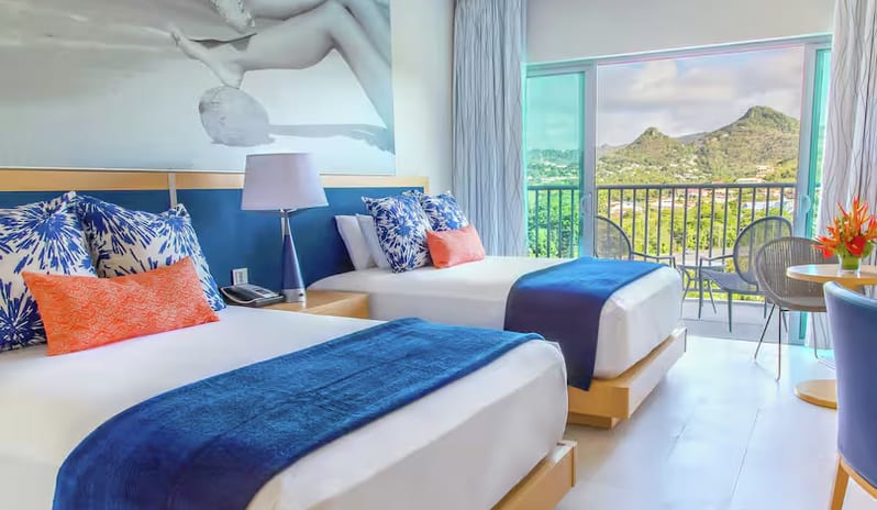 Mountain View Terrace Doubles two double beds with blue runner blankets, orange and patterned pillows, large wall art above the headboard, and open sliding glass doors leading to a balcony with a scenic mountain view at Harbor Club St. Lucia, Curio Collection by Hilton by Kenwood Travel