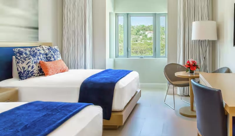 Island View Doubles two double beds with blue runner blankets, blue and white patterned pillows, a small wooden seating table, a desk, and a window opening to a lush green island view at Harbor Club St. Lucia, Curio Collection by Hilton by Kenwood Travel