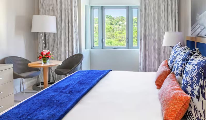 Island View King king bed, blue runner blanket, patterned pillows, a small seating area, and a window showcasing a green island view at Harbor Club St. Lucia, Curio Collection by Hilton by Kenwood Travel