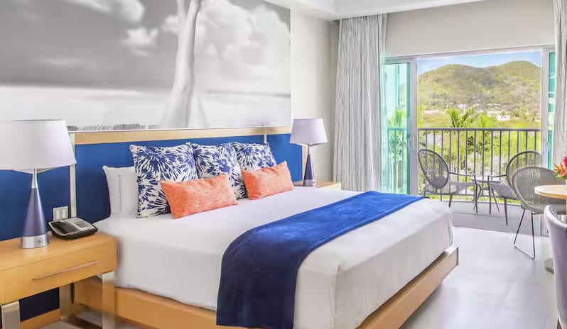 Harbor Club St. Lucia, Curio Collection by Hilton Marina View Terrace King king bed, blue runner blanket, orange accent pillows, and open sliding glass doors leading to a private balcony terrace with patio chairs overlooking a scenic landscape-Kenwood Travel