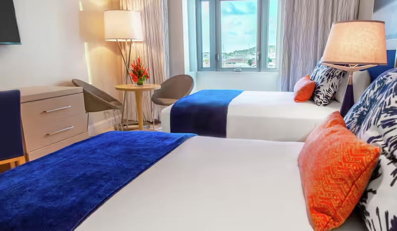 Marina View Doubles two beds with white linens, blue throws, and orange accent pillows, a small seating area, and a window with a city view at Harbor Club St. Lucia, Curio Collection by Hilton by Kenwood Travel