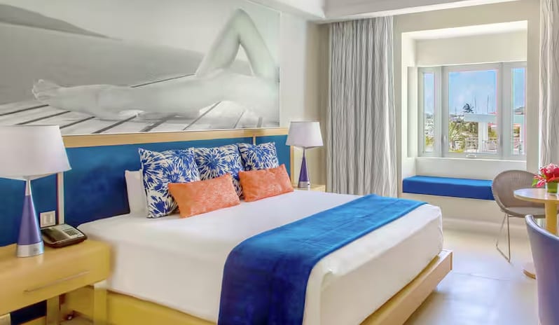 Marina View King rking bed with a blue runner blanket, orange and patterned blue pillows, a large black-and-white wall art piece, and a window with a blue cushioned window seat overlooking the marina at Harbor Club St. Lucia, Curio Collection by Hilton by Kenwood Travel
