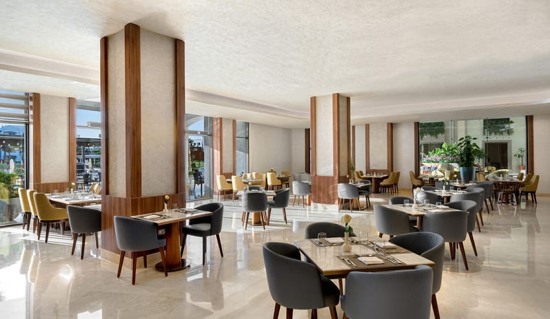 Veranda dining room showcasing contemporary square wooden tables, comfortable plush armchairs, and bright, sunlit interiors at Rixos Radamis Sharm El Sheikh by Kenwood Travel