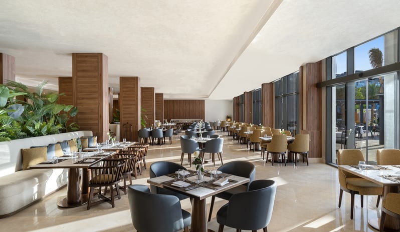 Veranda dining room showcasing contemporary square wooden tables, comfortable plush armchairs, and bright, sunlit interiors at Rixos Radamis Sharm El Sheikh by Kenwood Travel