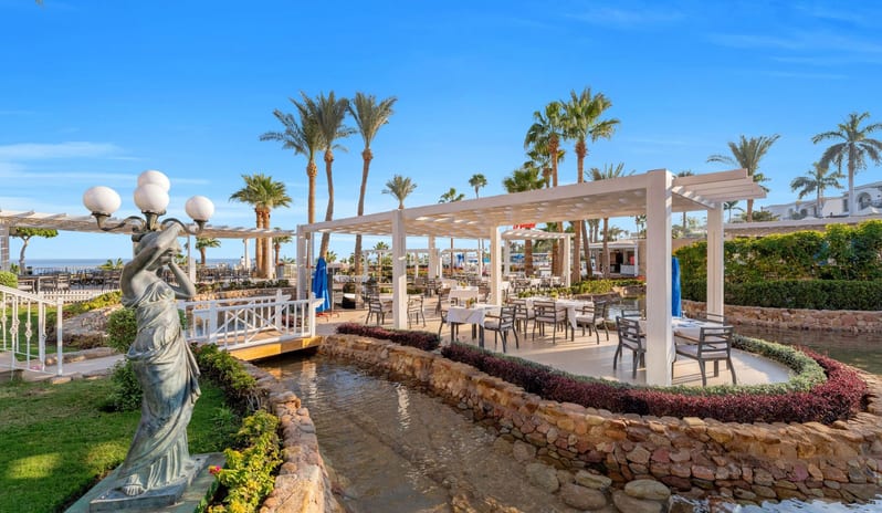 Adalar white pergolas, dining tables next to a stone-lined stream, a classic statue, and palm trees under a blue sky at Savoy Sharm El Sheikh by Kenwood Travel