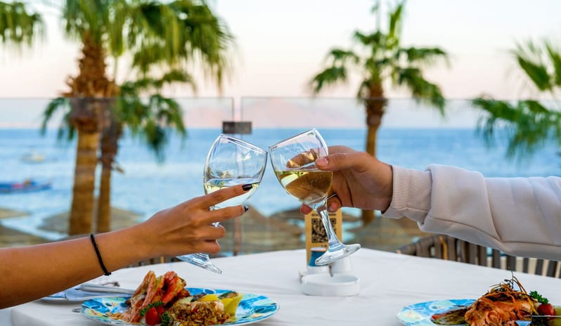 Seafood Island Close-up of a couple clinking white wine glasses over a seafood dinner at an outdoor oceanfront restaurant with palm trees at Savoy Sharm El Sheikh by Kenwood Travel