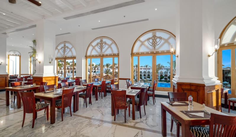 Tirana dining room featuring marble flooring, wooden tables, and large arched glass doors overlooking an outdoor swimming pool at Savoy Sharm El Sheikh by Kenwood Travel