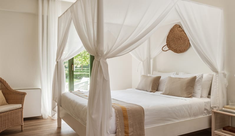 Family Interconnecting Room Shaded Veranda Bright neutral bedroom interior showcasing a white canopy bed draped in sheer curtains, a wicker armchair, and a decorative woven basket wall hanging at Grecotel LUXME Costa Botanica by Kenwood Travel