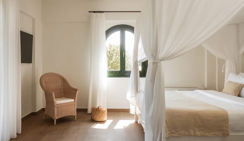 Family Summer Home Shaded Veranda & Gardens bedroom featuring a white four-poster canopy bed draped in sheer white fabric with a woven basket wall hanging at Grecotel LUXME Costa Botanica by Kenwood Travel