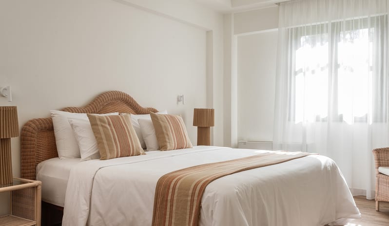 Family Interconnecting Room Shaded Veranda king bed, woven rattan headboard, striped accent pillows, and bright windows with sheer curtains at Grecotel LUXME Costa Botanica by Kenwood Travel