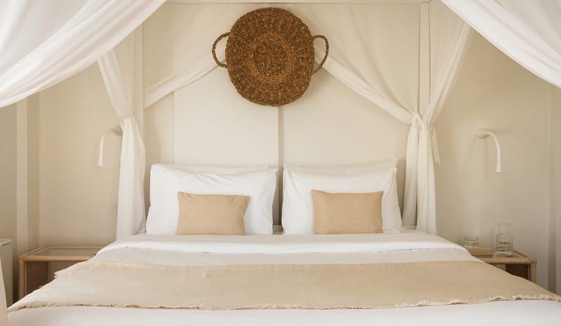 Family Summer Home Shaded Veranda & Gardens bedroom featuring a white four-poster canopy bed draped in sheer white fabric with a woven basket wall hanging at Grecotel LUXME Costa Botanica by Kenwood Travel