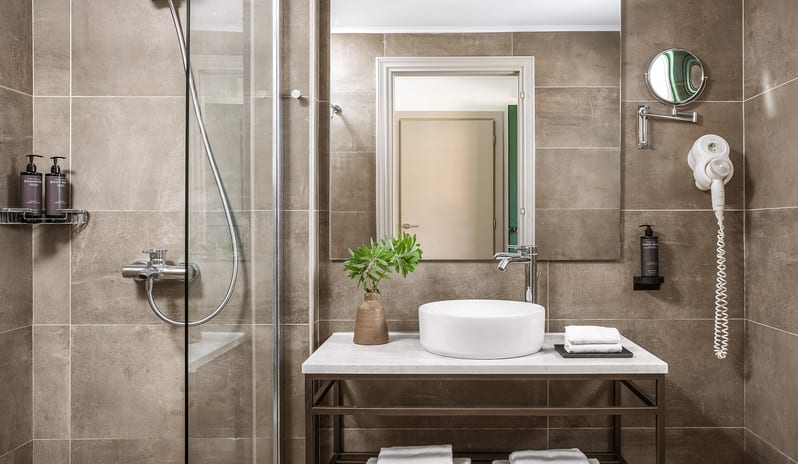 Family Interconnecting Room Shaded Veranda bathroom interior featuring warm brown wall tiles, an open metal console vanity with a marble top, and a sleek round ceramic vessel sink at Grecotel LUXME Costa Botanica by Kenwood Travel