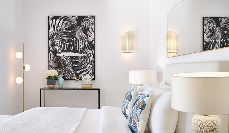 LUX ME Bungalow Poolside Close-up of a white resort bed with decorative pillows in front of a large black and white tropical leaf art piece at Grecotel LUXME Dama Dama by Kenwood Travel