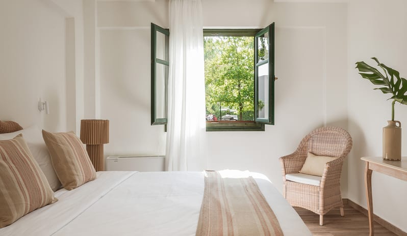 Family Interconnecting Room Shaded Veranda open green-trimmed windows with a garden view, a white bed with neutral striped linens, and a woven wicker armchair at Grecotel LUXME Costa Botanica by Kenwood Travel