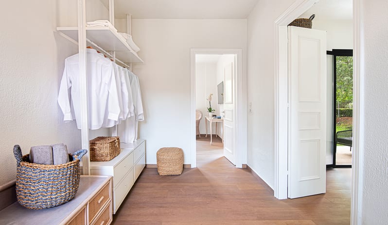 Family Summer Home Shaded Veranda & Gardens Minimalist open-frame wardrobe layout with white clothing rails, integrated low dressers, wood flooring, and woven rattan baskets for a coastal chic bohemian style at Grecotel LUXME Costa Botanica by Kenwood Travel