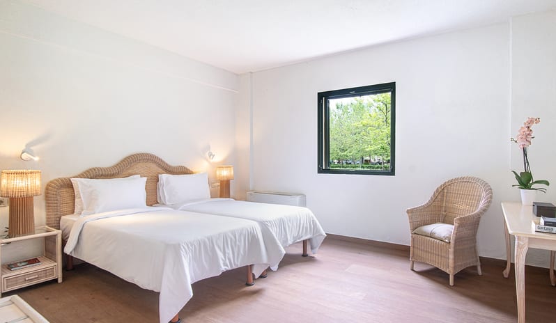 Family Summer Home Shaded Veranda & Gardens guest bedroom layout inside a luxury summer villa, showing twin beds with crisp white bedding, ample floor space, and a window framing an outdoor garden panorama at Grecotel LUXME Costa Botanica by Kenwood Travel