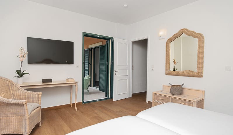 Family Summer Home Shaded Veranda & Gardens pacious twin bedroom layout in a family summer home featuring a wall-mounted flat-screen TV, a wicker armchair, and a decorative wave-edged mirror over a rattan chest of drawers at Grecotel LUXME Costa Botanica by Kenwood Travel