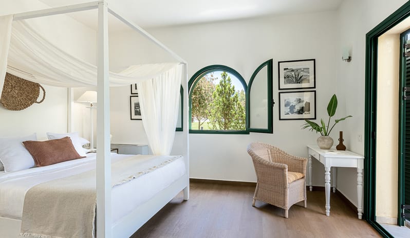 Family Summer Home Shaded Veranda & Gardens bedroom suite in a family summer home featuring a white four-poster canopy bed, an arched green window overlooking trees, a wicker armchair, and a small writing desk at Grecotel LUXME Costa Botanica by Kenwood Travel