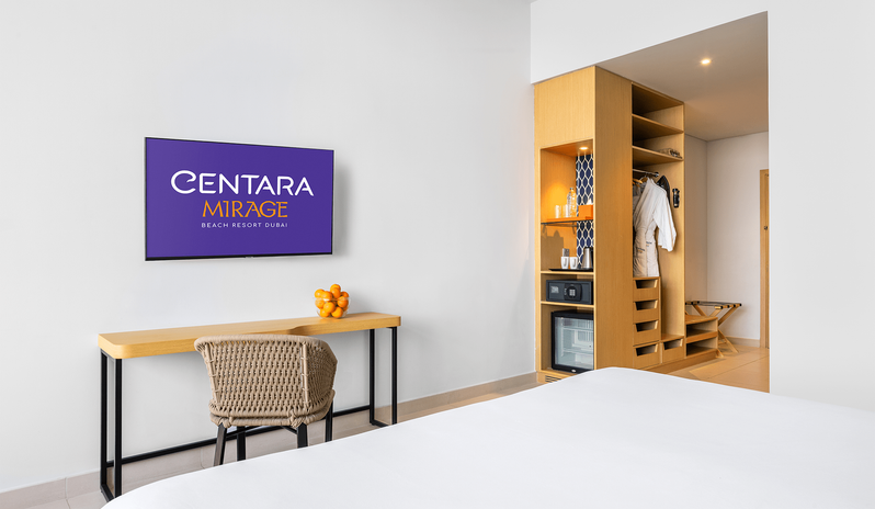 Superior Room King bed facing a wall-mounted TV, wooden desk with a bowl of oranges, and an open closet containing a safe, mini-bar, and bathrobes at Centara Mirage Beach Resort Dubai by Kenwood Travel