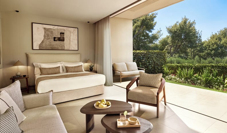 Double Room Garden View Luxury hotel suite with an open-concept bedroom, indoor seating area, and seamless glass doors opening to a private patio and lush garden at Sani Club by Kenwood Travel