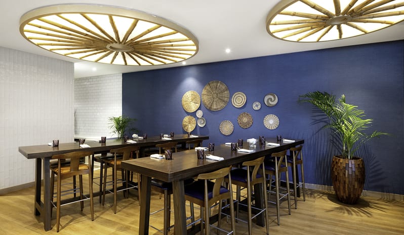 Suan Bua showcasing high-top communal dining tables with wooden chairs, warm lighting from unique cartwheel ceiling fixtures, and tropical potted plants against a blue wall at Centara Mirage Beach Resort Dubai by Kenwood Travel