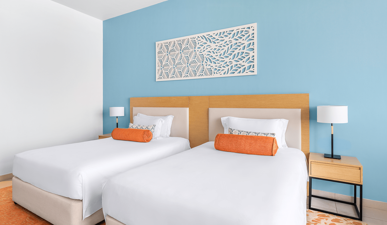 Family Room Panoramic Sea View Double Double room interior showing twin double beds, wooden headboard, bedside tables with lamps, and a bright blue feature wall at Centara Mirage Beach Resort Dubai by Kenwood Travel